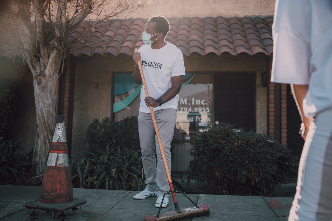 An adult volunteer cleaning a sidewalk with a broom, wearing a face mask for safety.